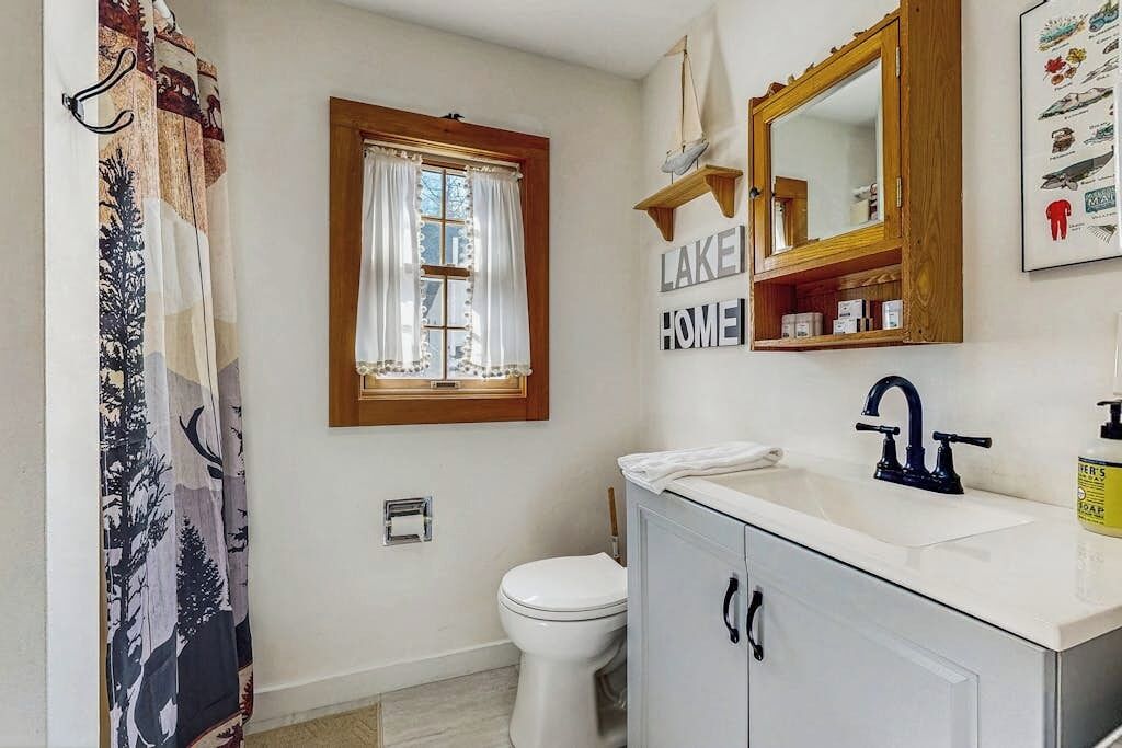 A bathroom with a white sink, a mirror, a window with curtains, and a toilet.