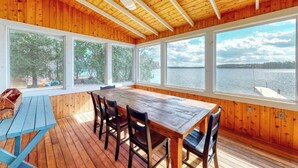 A wooden cabin with a large dining table, four chairs, and a view of a lake. - Lakefront home with private dock, kayaks, firepit, grill, & sunroom (Monmouth)
