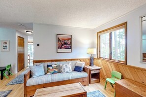 Ein gemütliches Zimmer mit einer Holzsofa, einem kleinen Tisch, einem Sessel, einer Lampe und einem großen Fenster.
