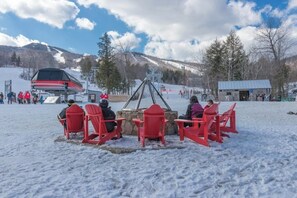 Eine Gruppe von Personen sitzt auf roten Stühlen um ein Steinkaminfeuer in einer verschneiten Winterlandschaft, im Hintergrund ist eine Seilbahn und Berge zu sehen.