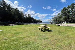 Un área de parque al aire libre con mesas de picnic, un campo cubierto de césped y un cuerpo de agua al fondo.