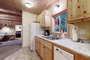 A compact kitchen with wooden cabinets, a built-in dishwasher, a refrigerator, and a window with a view of trees.
