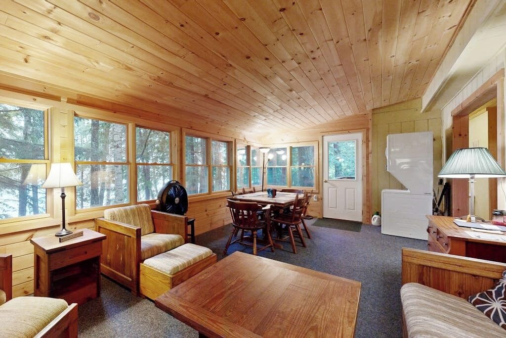 A cabin with wooden walls and ceiling, a dining area with a table and chairs, a sofa, and a refrigerator.