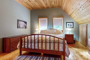 Ein Schlafzimmer mit Holzbalkendecke, zwei Fenstern mit Jalousien, einem Bett mit Holzrahmen, einem Nachttisch und einer Kommode.