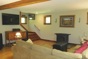 A living room with a sofa, a fireplace, a staircase, and a television mounted on the wall. - Spacious river home w/ pellet stove, outdoor dining area, fire pit, & BBQ grill (Litchfield)