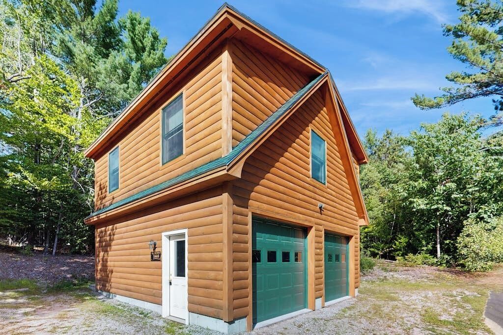 A two-story log cabin with a green garage door, surrounded by trees.