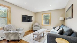 Un salon comprenant un canapé, un fauteuil, un banc et un téléviseur fixé au mur.