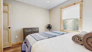 Une chambre à coucher avec un lit, une fenêtre avec des stores, un ventilateur, une commode et une porte.
