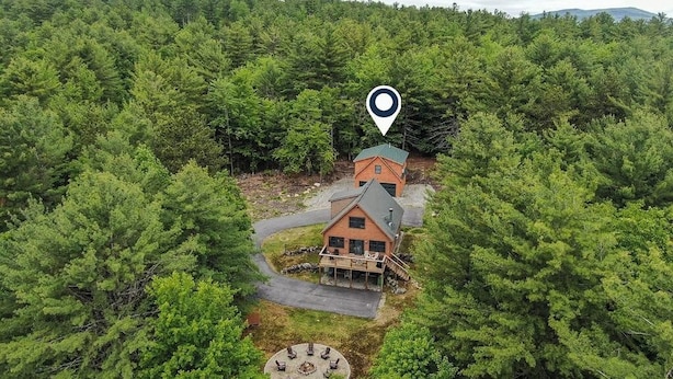 Un chalet entouré d’une forêt dense, doté d’un espace extérieur circulaire aménagé en salon et d’un escalier apparent menant au bâtiment principal.