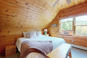 Ein Schlafzimmer einer Holzhütte mit Bett, Kissen, einem Nachttisch, einer Lampe und einem Fenster mit Blick.