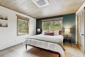 Ein Schlafzimmer mit einem Bett, zwei Fenstern, einem Nachttisch und einer Lampe.