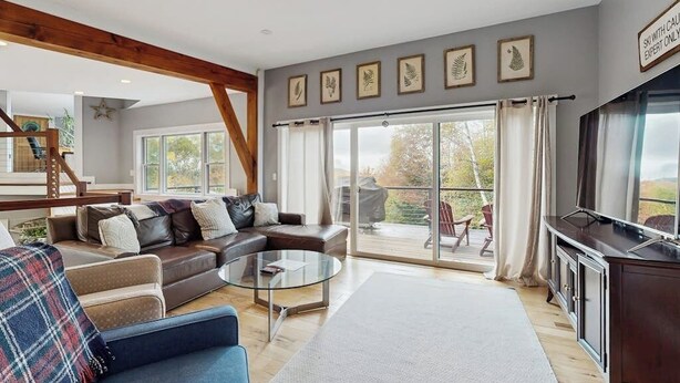 A spacious living room with a large sofa, a coffee table, and a TV. There is a balcony with a view of the outdoors. - Spacious Mountain Chalet with Sauna, Deck, and Ski/Snowboard Adventures Nearby (Bethel)