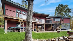 A two-story building with a covered patio area, a staircase, and a tree in the foreground. - Spacious Mountain Chalet with Sauna, Deck, and Ski/Snowboard Adventures Nearby (Bethel)