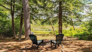 Two chairs facing a fire pit in a forested area with trees and a view of a field. - Lake Cabin | Fire Pit | Direct Lake Access | Pool | Near Ski Area (Bridgton)