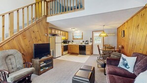 A spacious living area with a wooden staircase, a television, a sofa, and a dining table. - Lake Cabin | Fire Pit | Direct Lake Access | Pool | Near Ski Area (Bridgton)