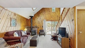 A cozy loft-style living room with a fireplace, wooden walls, a TV, and a staircase leading to a balcony. - Lake Cabin | Fire Pit | Direct Lake Access | Pool | Near Ski Area (Bridgton)