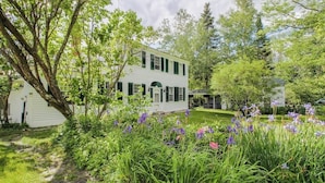 A two-story white house with green shutters, surrounded by lush greenery and flowering plants. - Historic home with porch & mountain/valley views - great for weddings (Paris)
