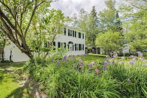 A two-story white house with green shutters, surrounded by lush greenery and flowering plants. - Historic home with porch & mountain/valley views - great for weddings (Paris)