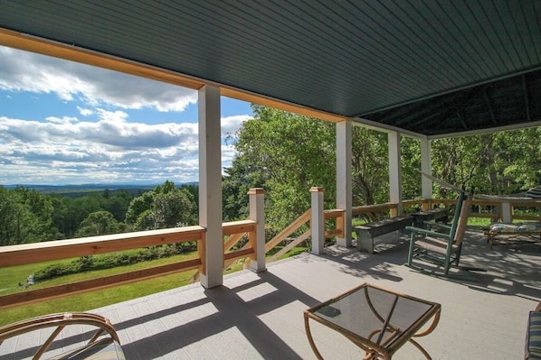 A covered outdoor deck with wooden railings, seating, and a view of a forested landscape. - Historic home with porch & mountain/valley views - great for weddings (Paris)