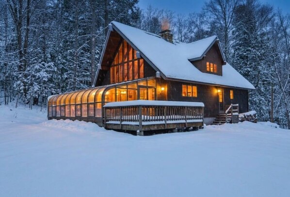 A wooden cabin with a large glass windowed porch, surrounded by snow-covered trees.