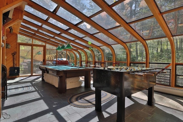 A spacious indoor area with a pool table, foosball table, and large windows offering views of a forested area.