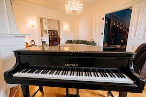 Un piano de cola en una habitación amplia con candelabro, escalera y vistas a una biblioteca.
