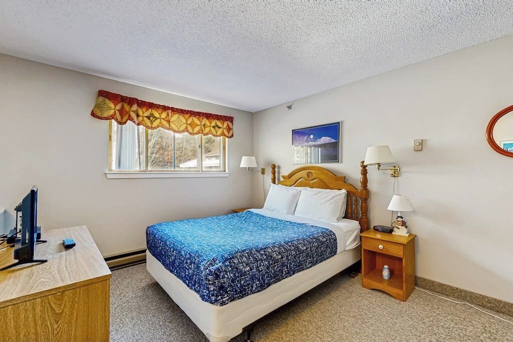 A bedroom with a bed, bedside tables, a desk, a window with curtains, a wall-mounted lamp, and a framed picture.