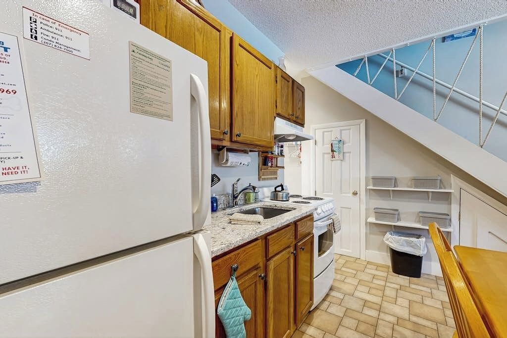 Una cocina con armarios de madera, un refrigerador blanco, una estufa y un lavamanos. Hay una ventana sobre el lavamanos y una puerta que conduce a otra habitación.