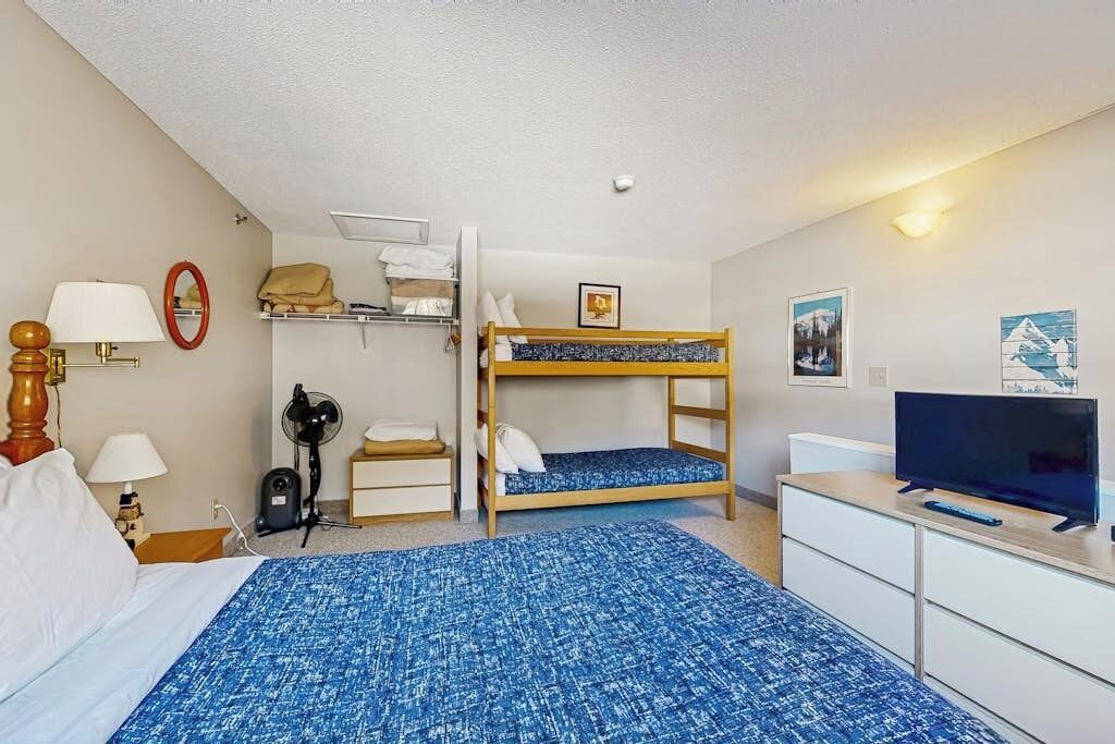 A hotel room with a bunk bed, a desk with a television, a lamp, a fan, and a shelf with folded clothes.