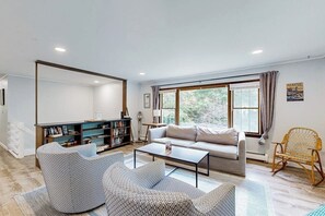 A living room with a sofa, two patterned chairs, a coffee table, a bookshelf, and a window with curtains.