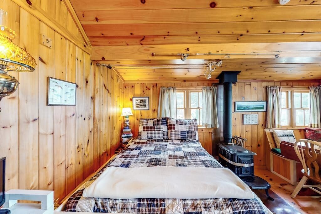 A wooden cabin room with a bed, a fireplace, a desk, and a window.