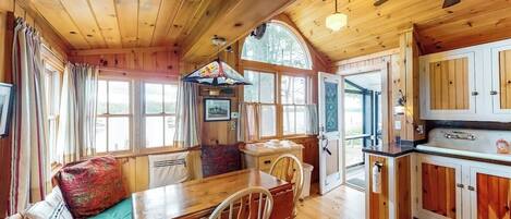 A wooden cabin interior with a dining area, kitchen, and a large window.