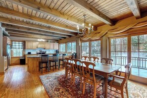 A spacious kitchen with wooden beams, a dining area with a large table and chairs, and large windows offering a view of the outdoors. - Expansive Retreat Near the Slopes with Media Room, Two Kitchens, Stone Fireplace (Newry)