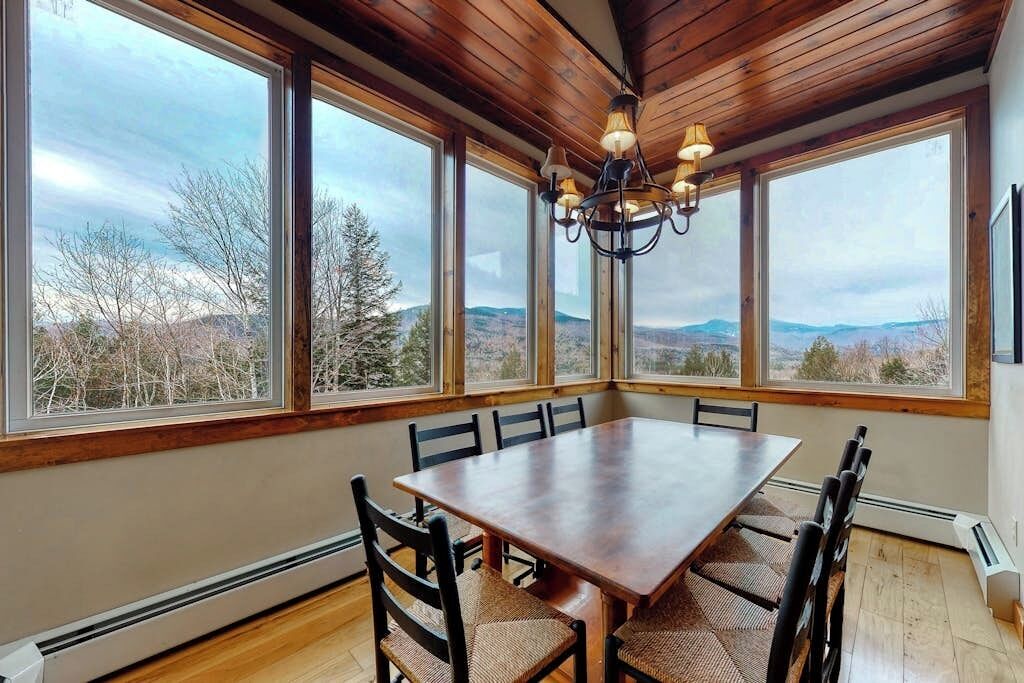 Un espace repas comprenant une grande table et des chaises, de grandes fenêtres donnant sur des arbres et des collines, et un lustre.