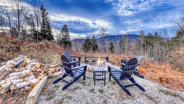 Ein Außenbereich mit Adirondack-Stühlen und einem Feuerkorb, umgeben von einer natürlichen Landschaft mit Bäumen und Bergen im Hintergrund.