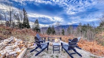 Un espace extérieur aménagé avec des chaises Adirondack et un foyer, entouré d’un paysage naturel avec des arbres et des montagnes en arrière-plan.