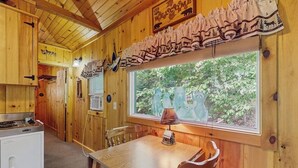 En stuga med träinredning, innehållande ett matbord, stolar, ett fönster med utsikt över grönskan, en spis och ett kylskåp.