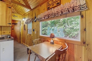 Ein Holzblockhaus-Interieur mit Esstisch, Stühlen, einem Fenster mit Blick auf Grünfläche, einem Herd und einem Kühlschrank.