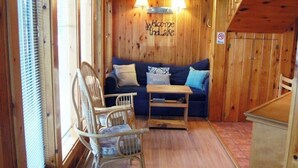 A cabin interior with a wooden wall, a blue sofa, a wooden table, and wicker chairs. - Dog-Friendly Lakefront Cottage w/Lake Access, Deck, Gas Grill, A/C (Standish)