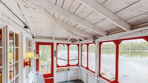 Una terraza techada con una mesa y sillas, ventanas amplias y vista a un lago tranquilo.