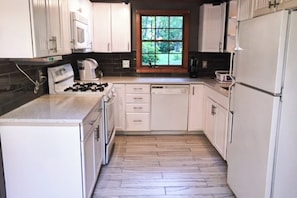A kitchen with white cabinets, a stove, a microwave, and a window with a view of greenery. - Private waterfront cabin with large deck, private dock, kayaks & gas grill (Naples)