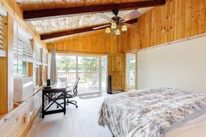 A room with a bed, a desk, and a chair, a ceiling fan, and large windows.