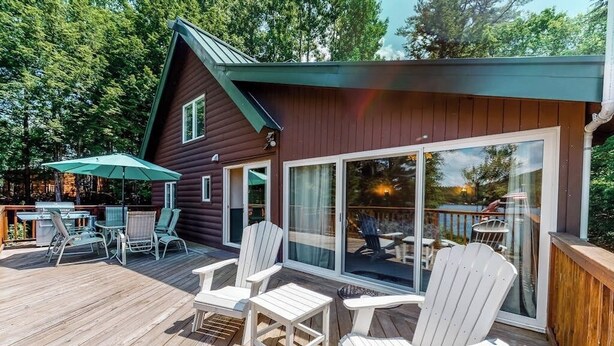 Um deck de madeira com cadeiras Adirondack e uma mesa, uma porta de vidro que dá para uma área de deck, e um pequeno edifício com telhado verde.