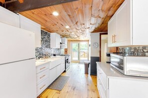 A modern kitchen with white cabinets, a stone backsplash, wooden ceiling beams, and a view to the outside.