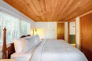 A bedroom with a wooden ceiling, a bed with white bedding, a nightstand, a lamp, and a window with blinds.
