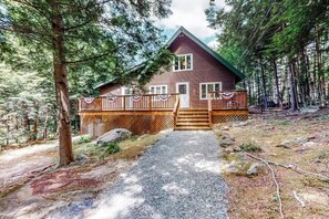 A cabin with a wooden deck, a gravel path leading to the entrance, and surrounded by dense forest.