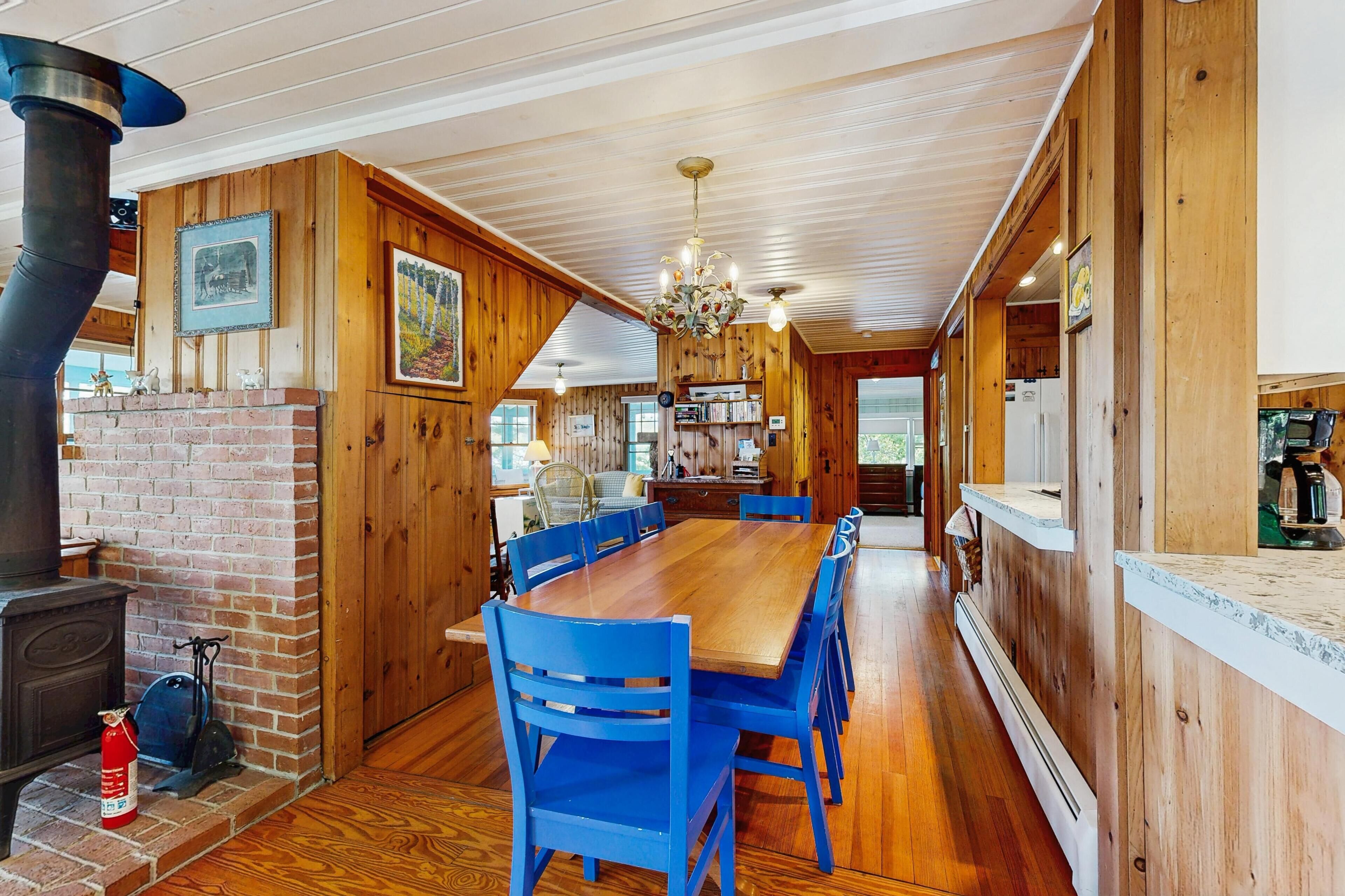 Un intérieur en bois avec une cheminée en briques, une table à manger, des chaises bleues et un coin cuisine.