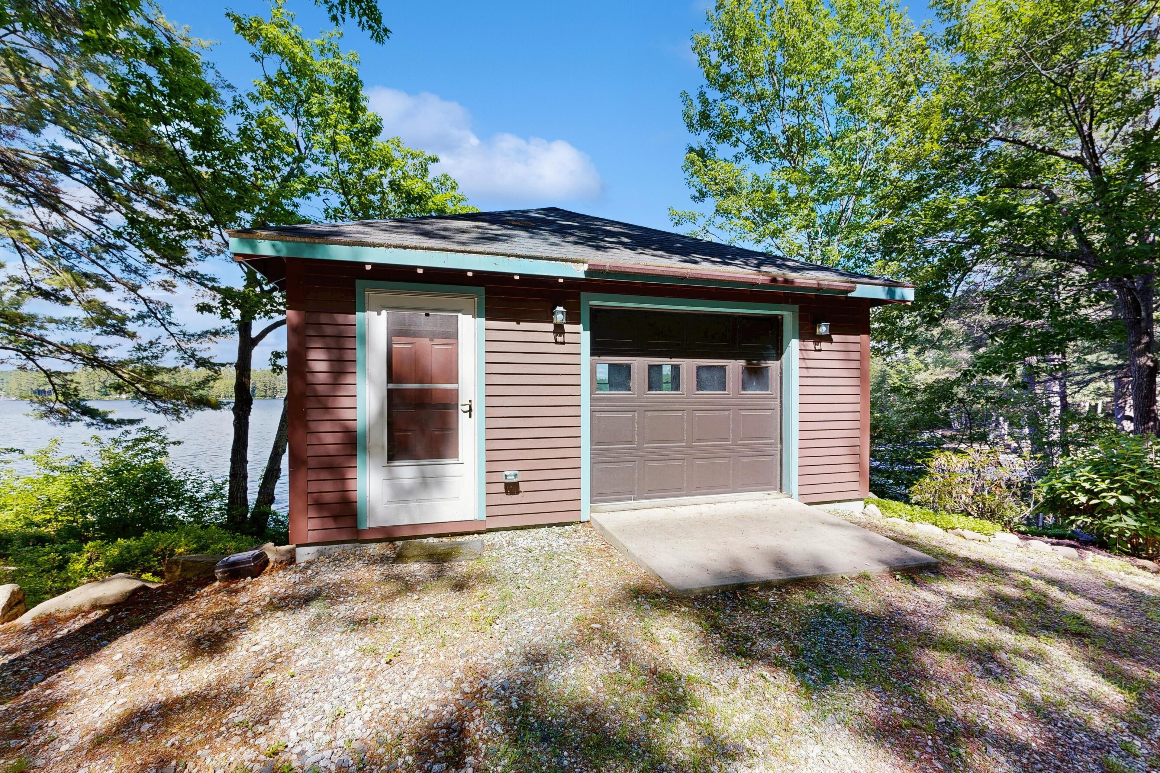 Une petite cabane avec une seule porte et un garage, entourée d’arbres et bordée par un lac.