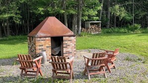 Ein gemauerter Außenkamin mit Schornstein, umgeben von Adirondack-Stühlen, in einer grünen Wiese mit Bäumen im Hintergrund.
