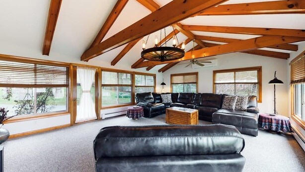 Ein geräumiges Wohnzimmer mit hoher Decke und freiliegenden Holzbalken, großen Fenstern und einem schwarzen Ledersofa.